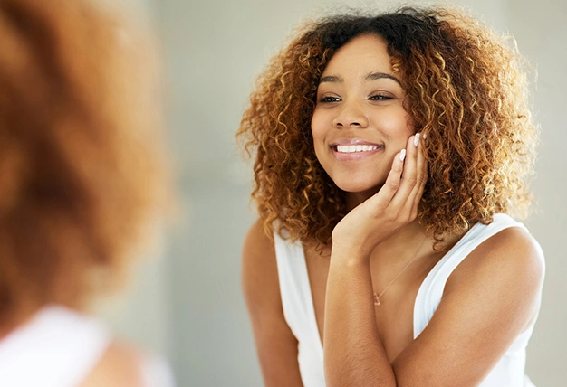 Woman smiling while touching her face in bathroom mirror, displaying healthy teeth and skin.